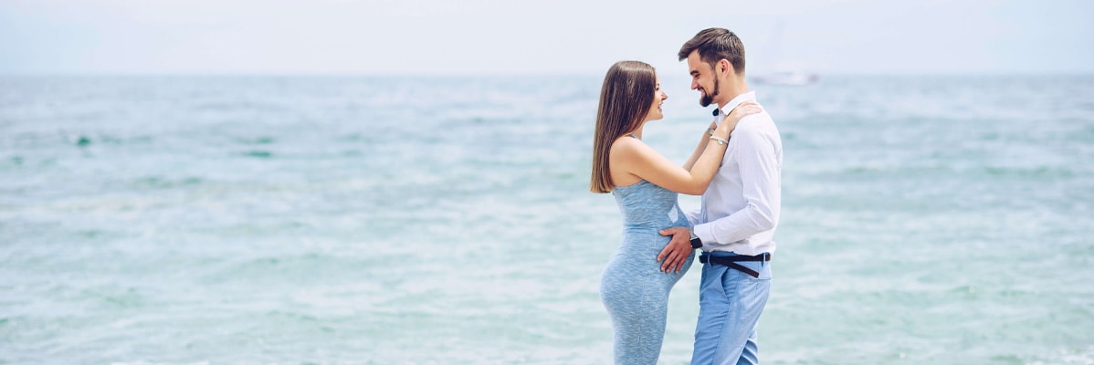 Donna incinta e uomo che si abbracciano vicino al mare.