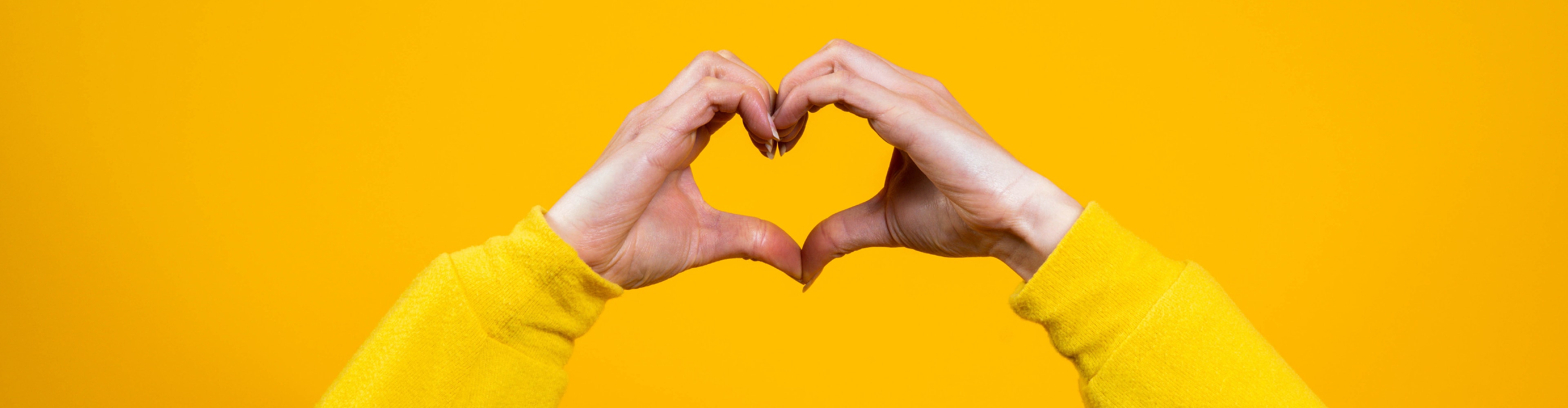 Hände formen ein Herz auf gelbem Hintergrund, Symbol für Liebe und Fürsorge.