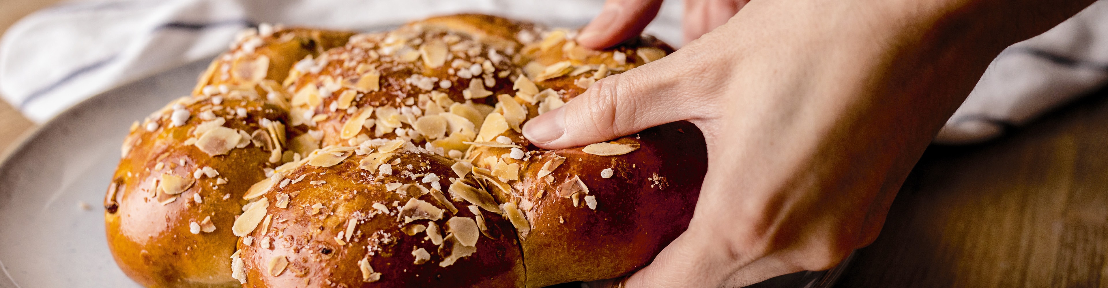 Hände greifen nach einem goldenen Zopfgebäck, garniert mit Mandelblättchen und Hagelzucker.