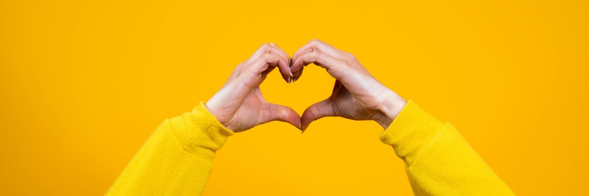Mani che formano un cuore su sfondo giallo, simbolo di amore e cura.