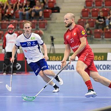 Due giocatori di floorball in azione su un campo indoor, uno con maglia bianca e blu, l'altro rossa.