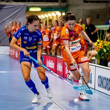 Due donne giocano a floorball con mazze e palla in un campo sportivo.