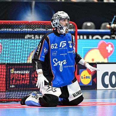 Un portiere di floorball in ginocchio in campo, con una rete da porta e sponsor sullo sfondo.