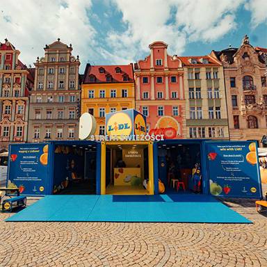 Ein blauer und gelber Lidl-Stand mit frischem Obst und Gemüse vor farbigen Gebäuden.
