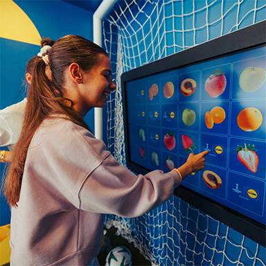 Frau spielt ein interaktives Spiel mit Fruchtbildern auf einem großen Bildschirm in einem Fußballzimmer.