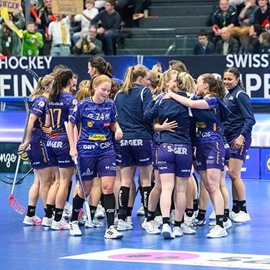Squadra di floorball femminile in campo, con spettatori e sponsor sullo sfondo.