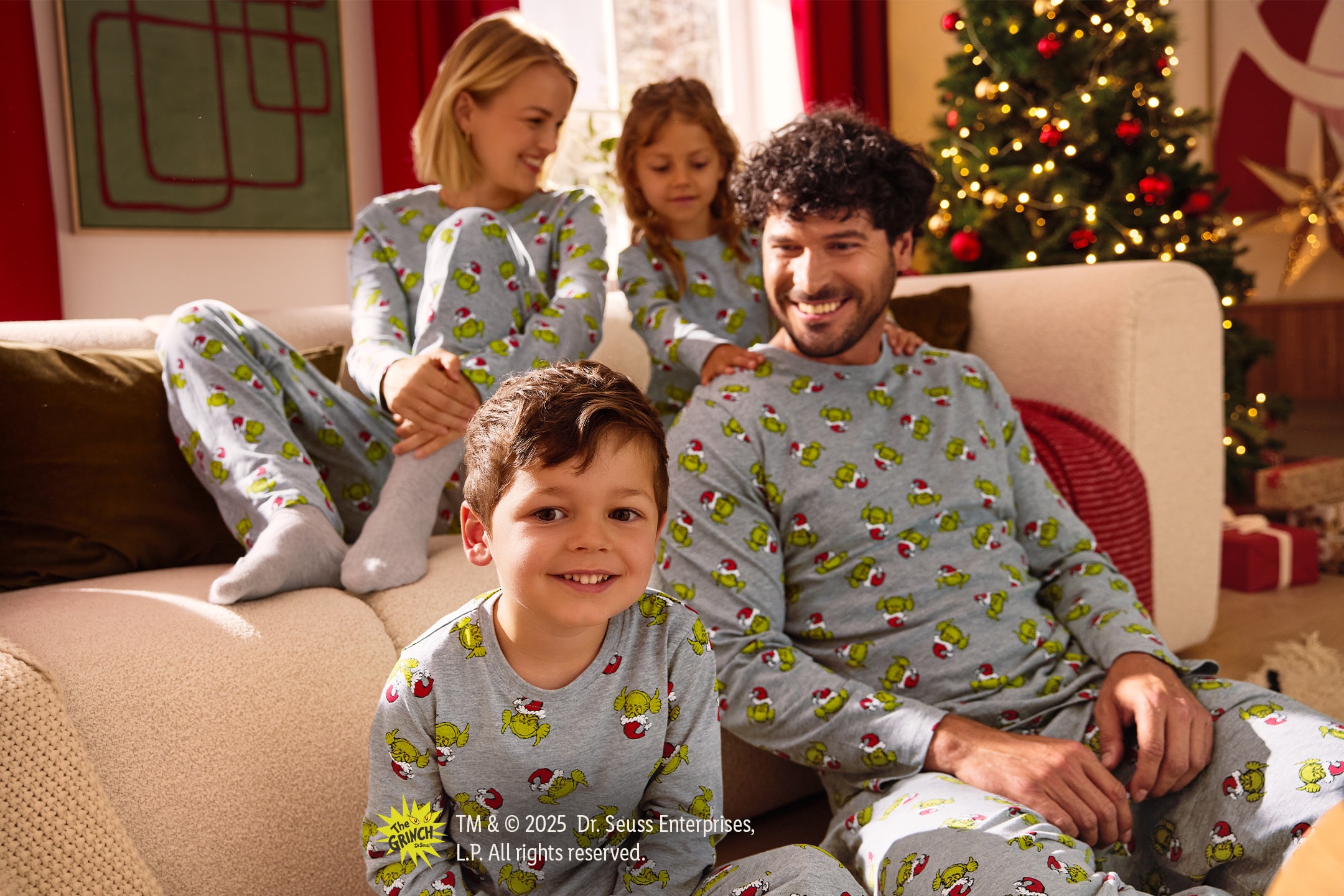 Familie in Grinch-Weihnachtspyjamas, auf einem Sofa neben einem Weihnachtsbaum sitzend.