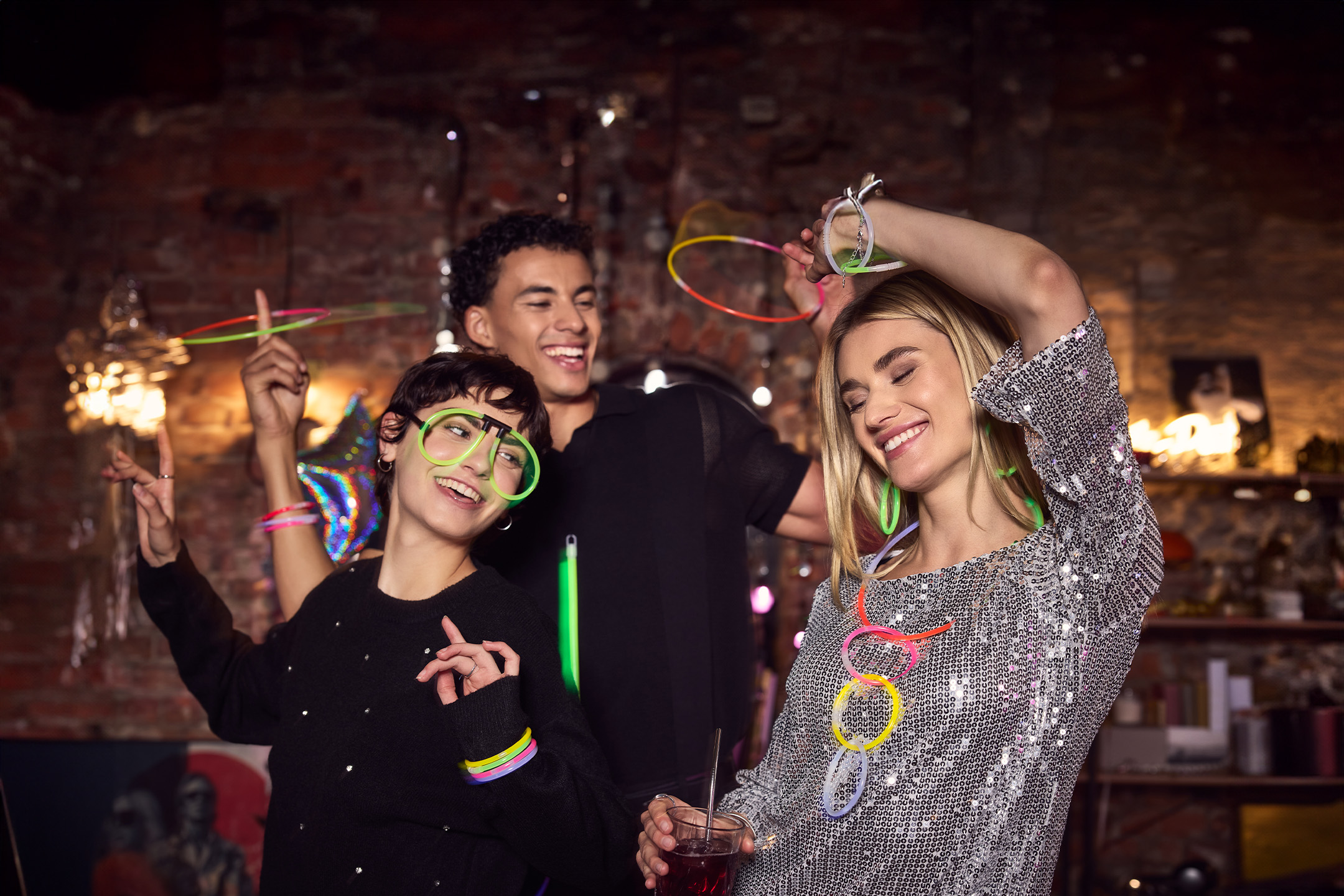Drei lächelnde Freunde tanzen auf einer Party und tragen leuchtende Accessoires.