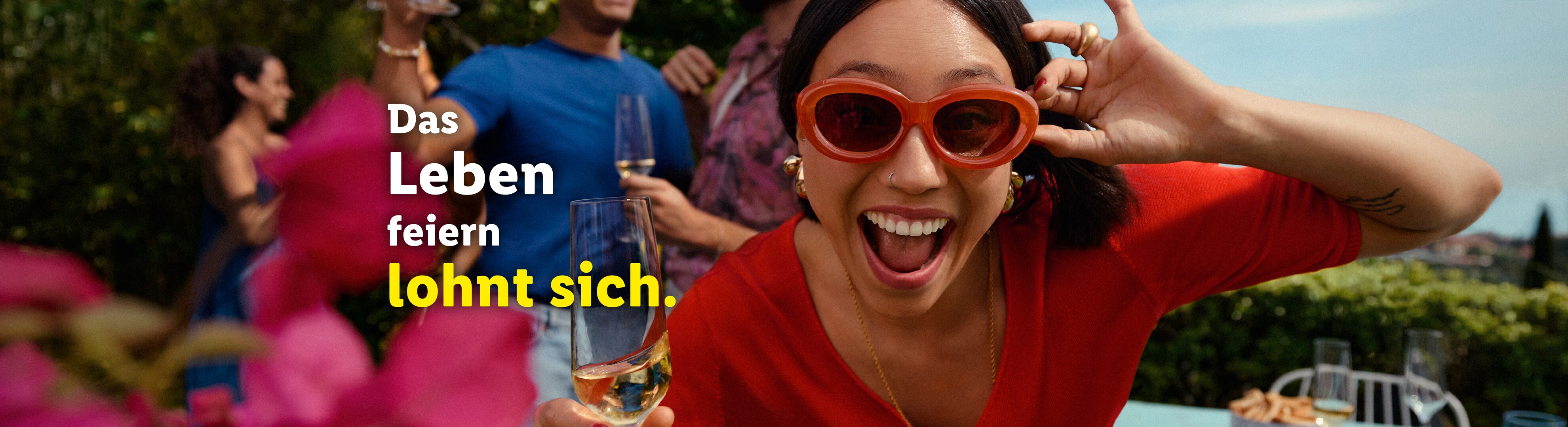 Eine lächelnde Frau mit orangefarbener Sonnenbrille hält ein Weinglas auf einer Gartenparty.