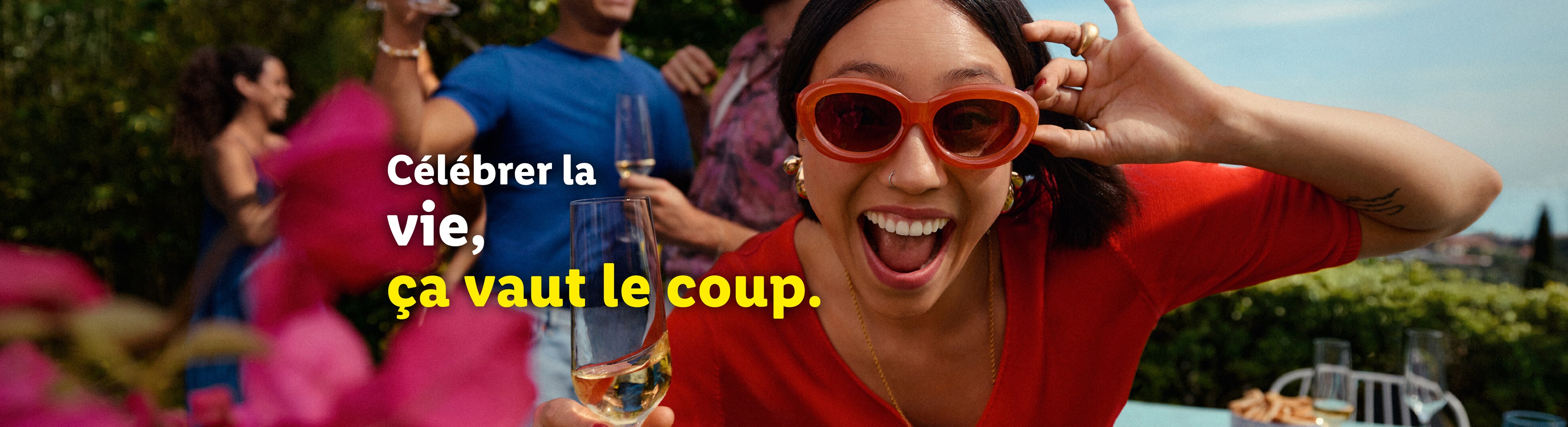 Une femme souriante avec des lunettes de soleil rouges tient un verre de vin, avec des amis en arrière-plan.
