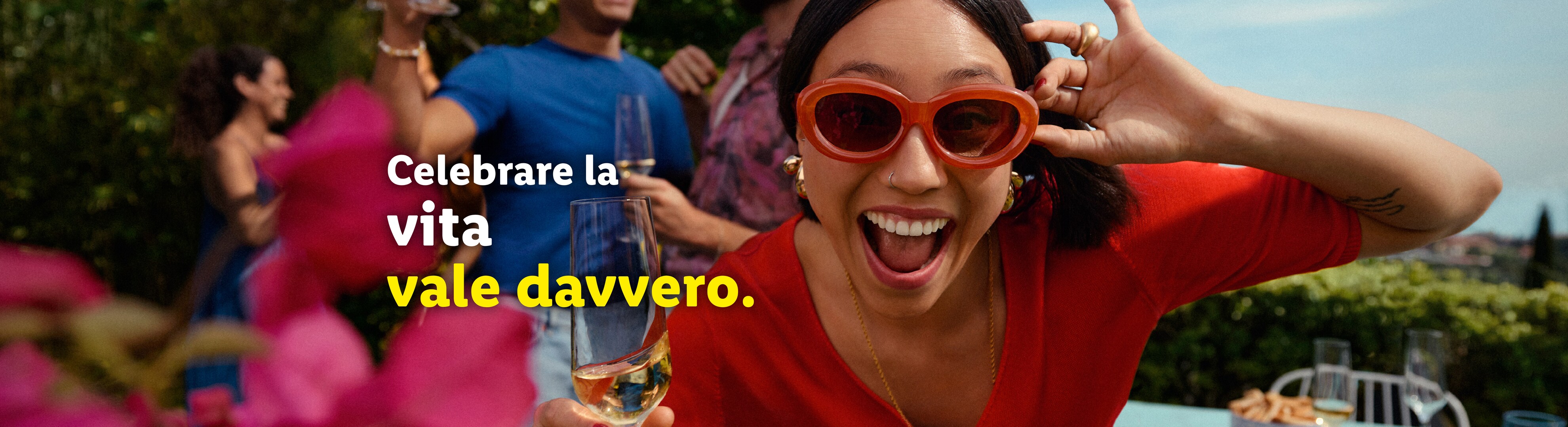 Una donna sorridente con occhiali da sole rossi tiene un bicchiere di vino bianco a una festa.