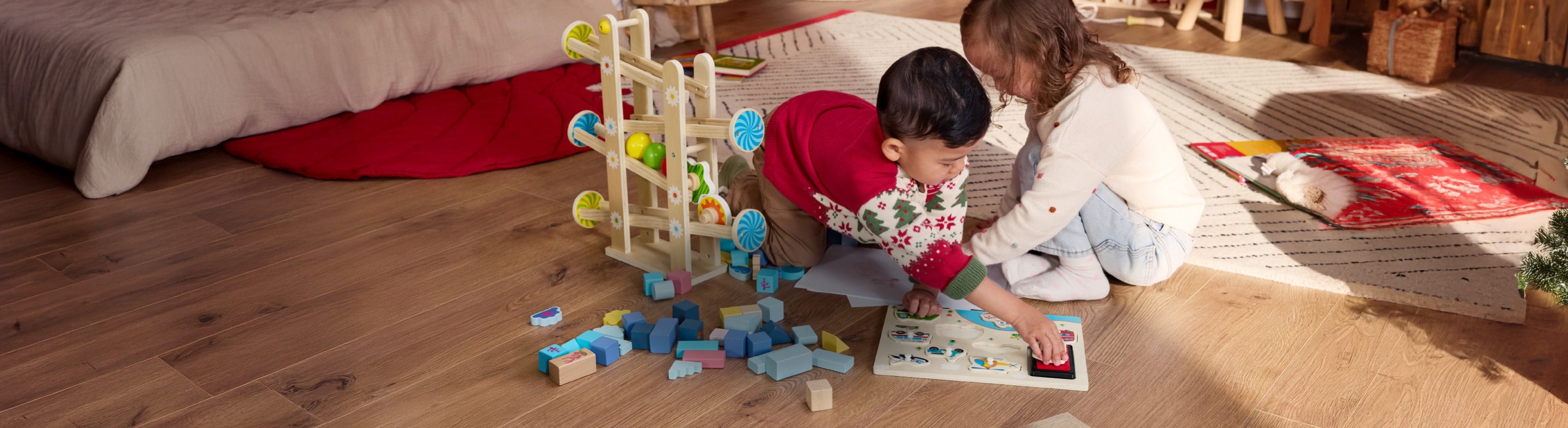 Zwei Kinder spielen mit einer Holzkugelbahn und Bauklötzen auf dem Boden.