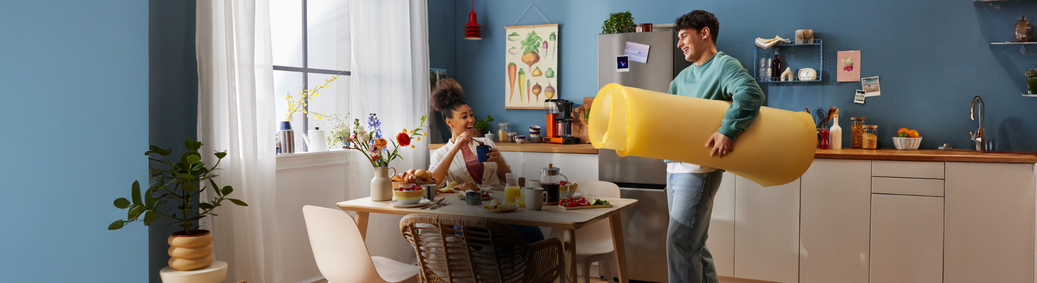 Un uomo trasporta un grande galleggiante giallo mentre una donna fa colazione a tavola in una cucina luminosa.