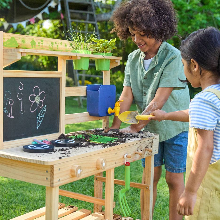 Deux enfants jouent avec une cuisine de boue en bois, avec un tableau noir et des plantes en pot.