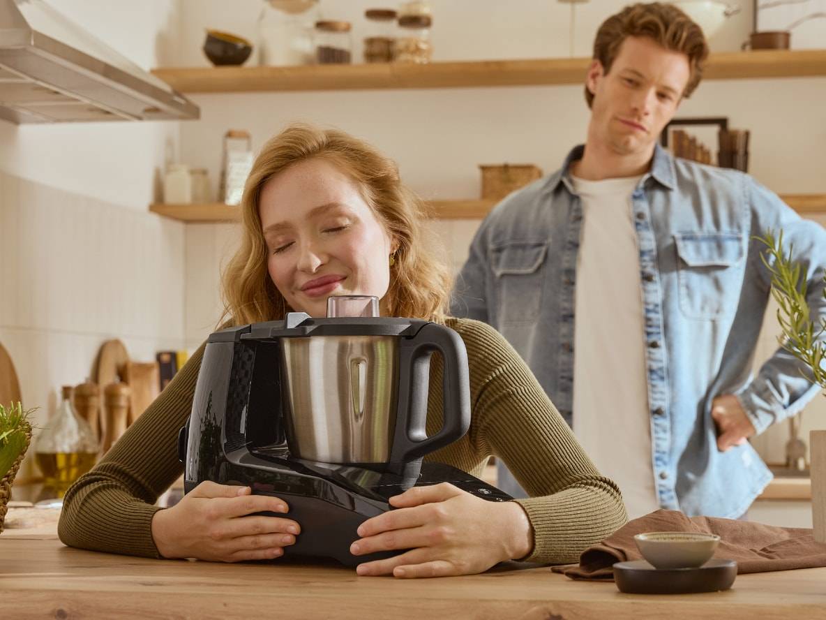 Eine Frau umarmt eine Monsieur Cuisine Küchenmaschine, ein Mann schaut zweifelnd zu.