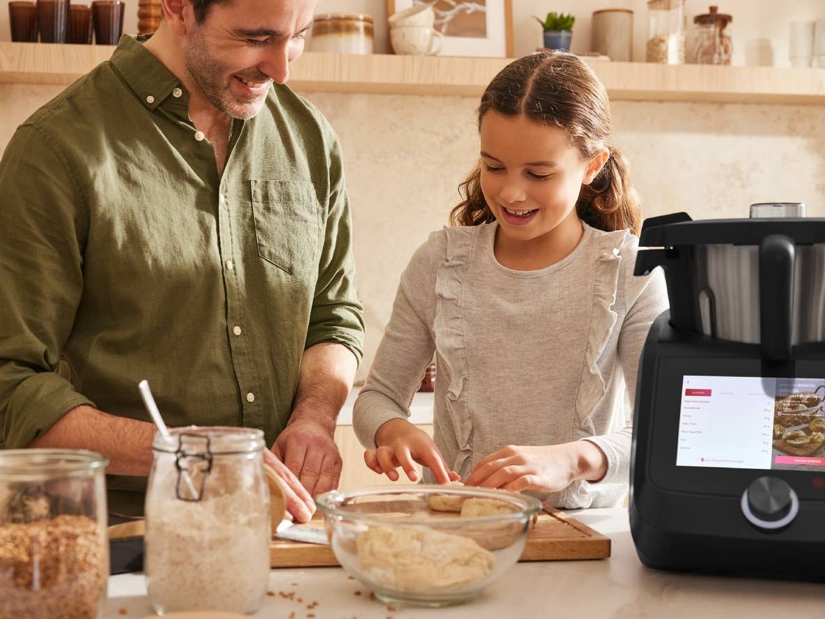 Lächelnder Vater und Tochter bereiten Teig mit einer Monsieur Cuisine Küchenmaschine zu.
