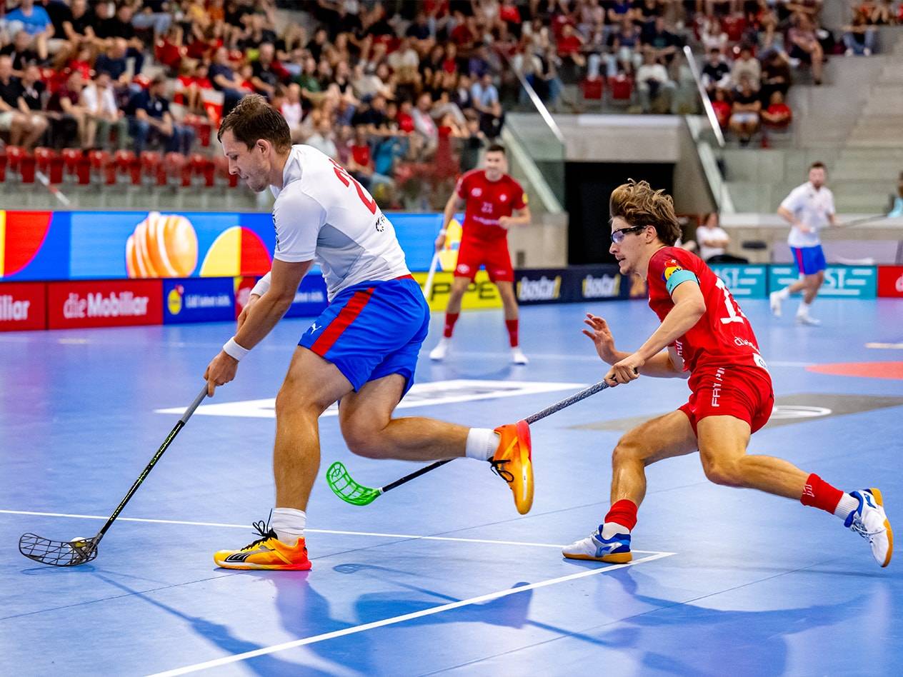 Due giocatori di floorball in azione su un campo blu, con spettatori sullo sfondo.