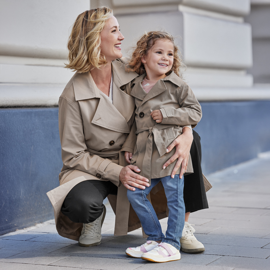Deux femmes en trench-coats beiges, une adulte et une enfant, souriant à l'extérieur.