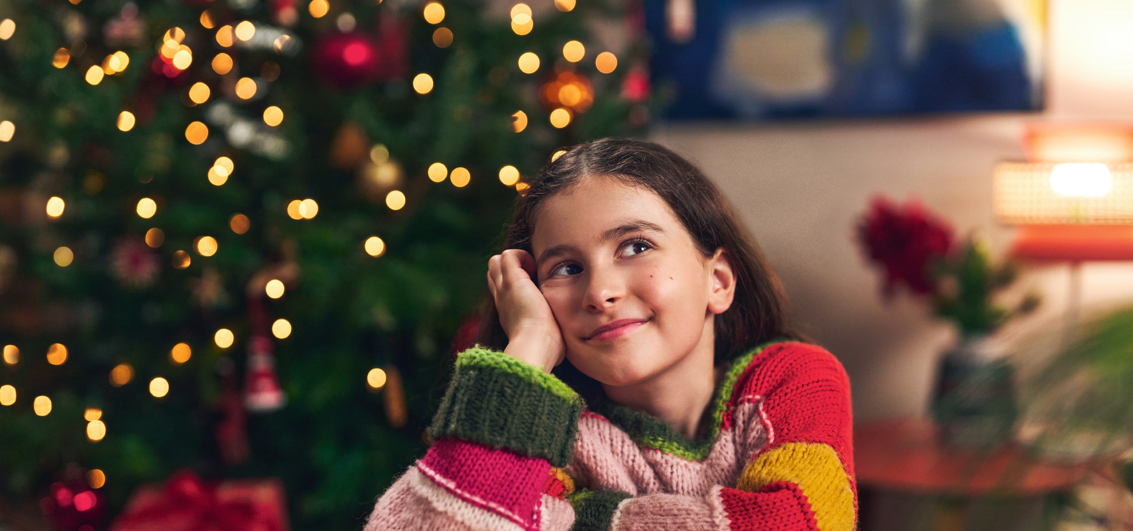 Lächelndes Mädchen in buntem Pullover, beleuchteter Weihnachtsbaum im Hintergrund.