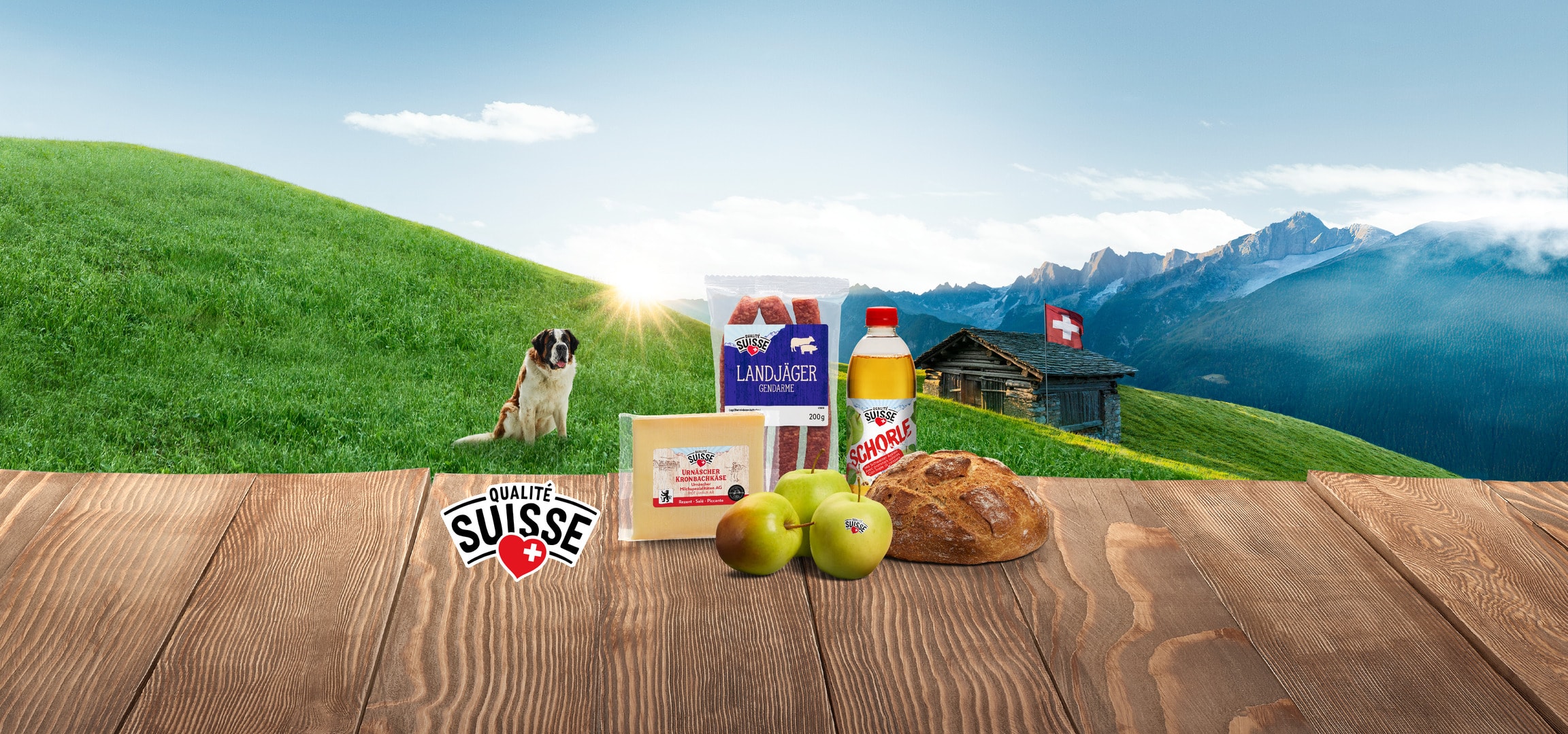 Schweizer Lebensmittel wie Käse, Wurst, Brot und Äpfel auf einem Holztisch mit einem Bernhardiner und Alpenlandschaft.