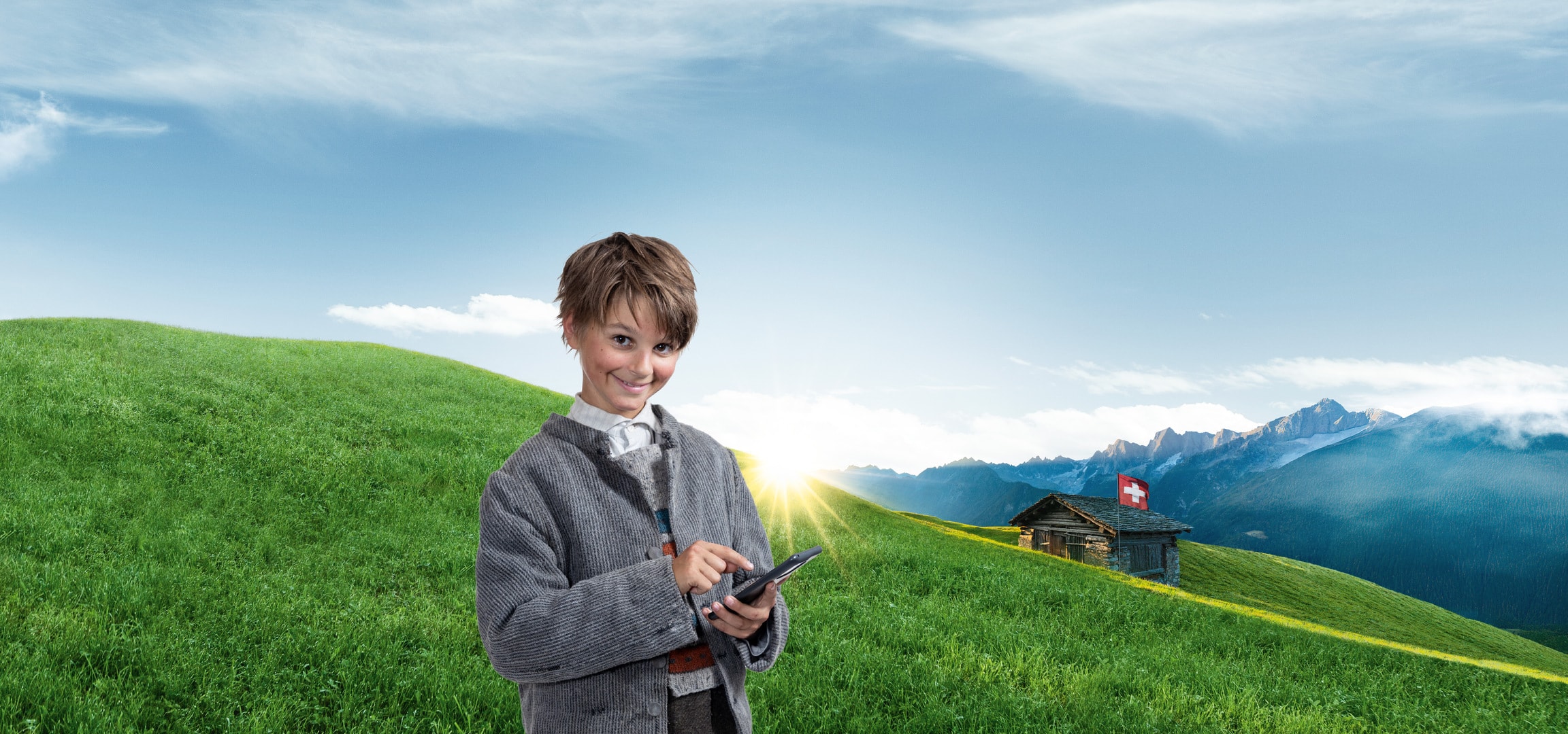 Ein lächelnder Junge benutzt ein Tablet in einer Schweizer Berglandschaft mit einer Hütte.