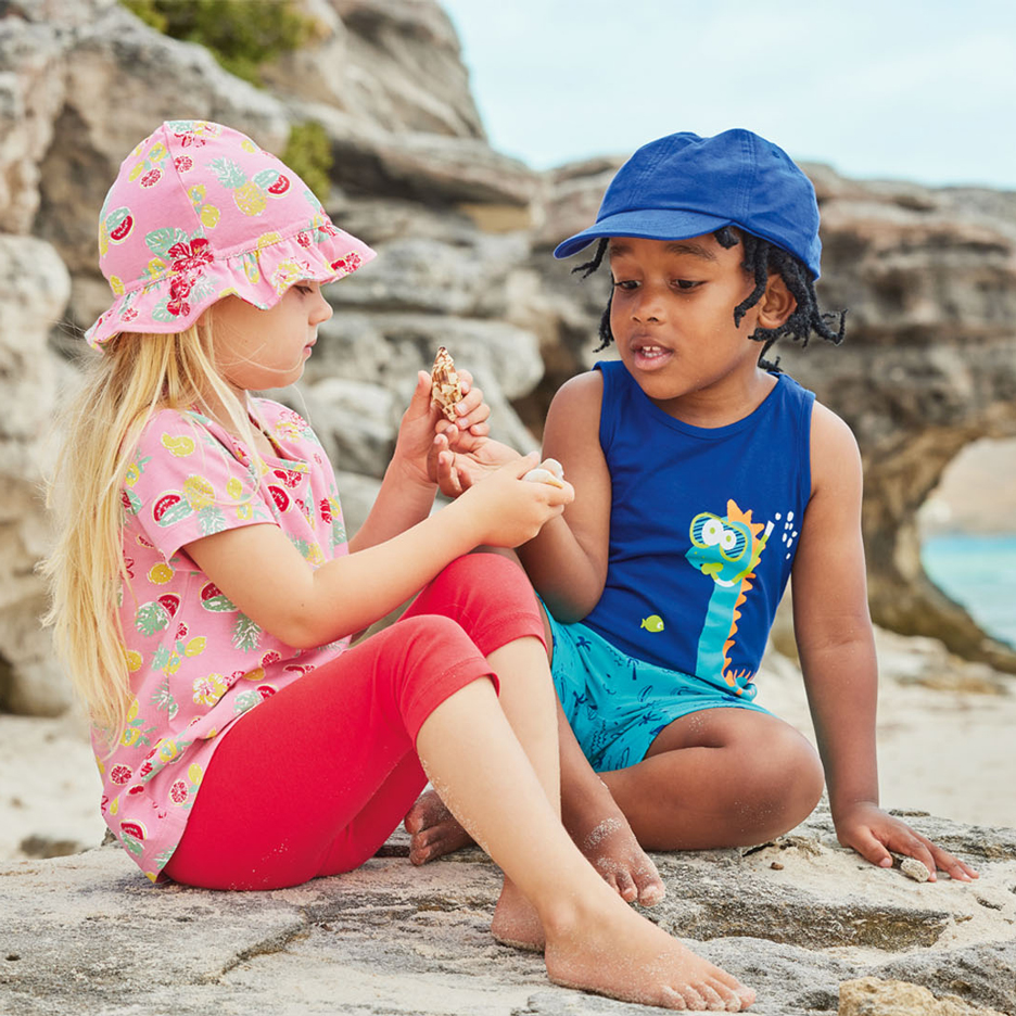 Deux enfants sur une plage, l'un avec un chapeau de soleil rose et un haut à motifs de fruits, l'autre avec une casquette bleue et un débardeur dinosaure, tenant des coquillages.