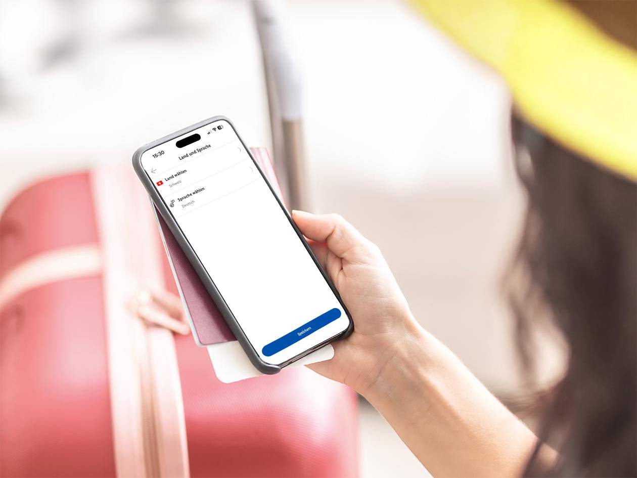 Frau hält Smartphone mit Sprachauswahl und Reisepass vor rotem Koffer.