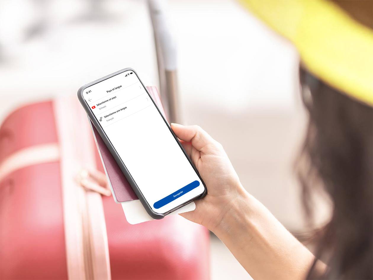 Une personne tenant un smartphone affichant les options de pays et de langue, avec un passeport et une valise en arrière-plan.