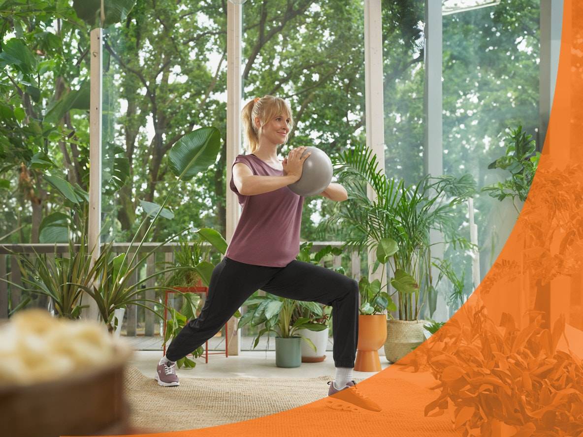Frau macht Ausfallschritte mit einem Medizinball in einem hellen Raum voller Pflanzen.