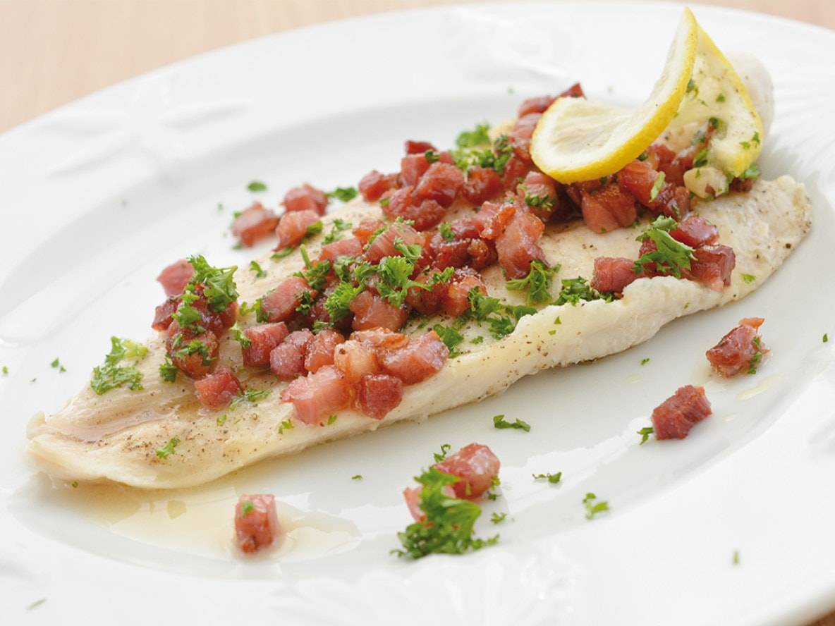 Gebratenes Fischfilet mit Speckwürfeln, Petersilie und Zitrone auf einem weißen Teller.