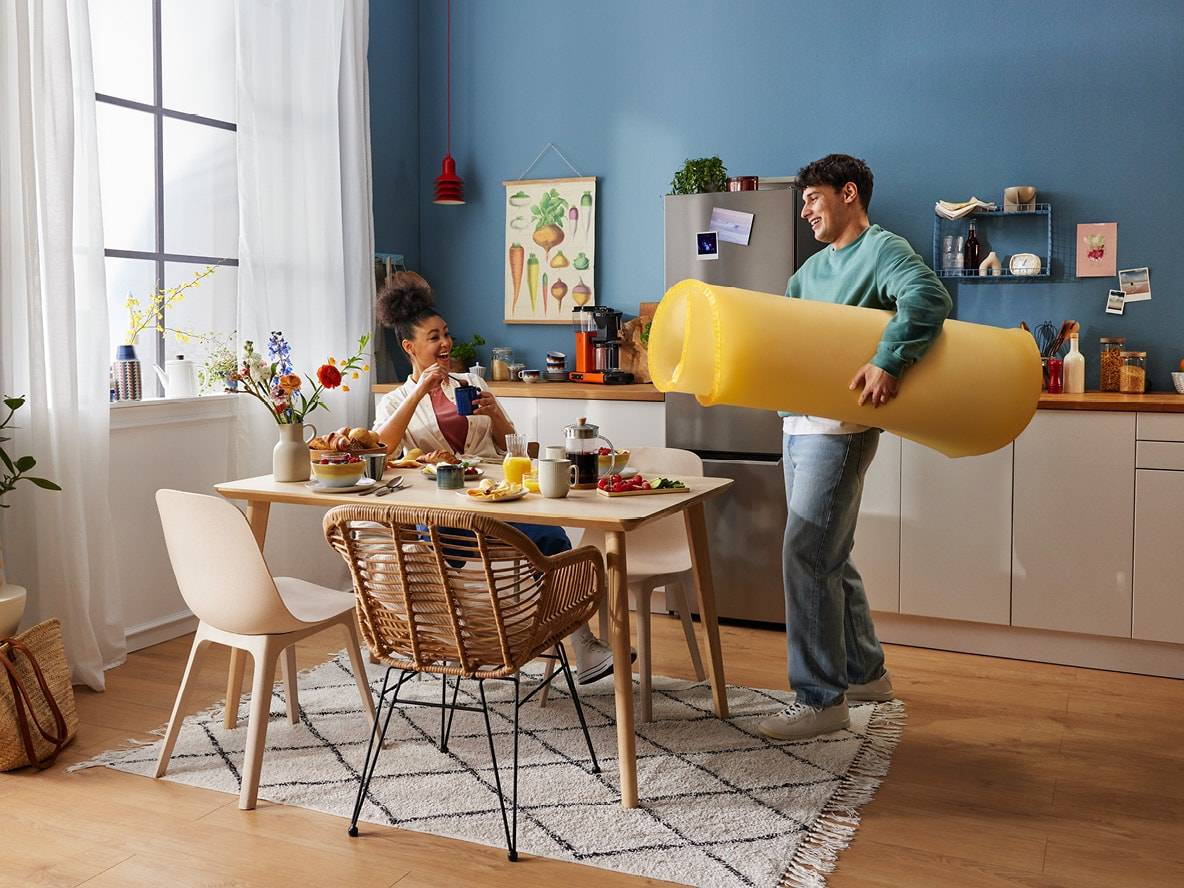 Ein Paar in einer Küche, die Frau frühstückt, der Mann trägt eine riesige Nudel.