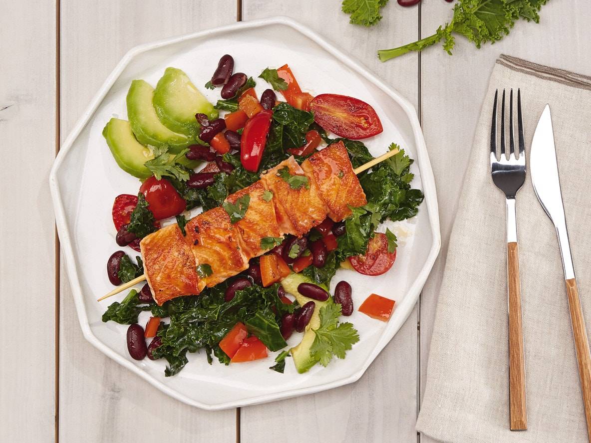 Lachsspieß mit Grünkohl, Kidneybohnen, Avocado und Tomaten auf einem weißen Teller.