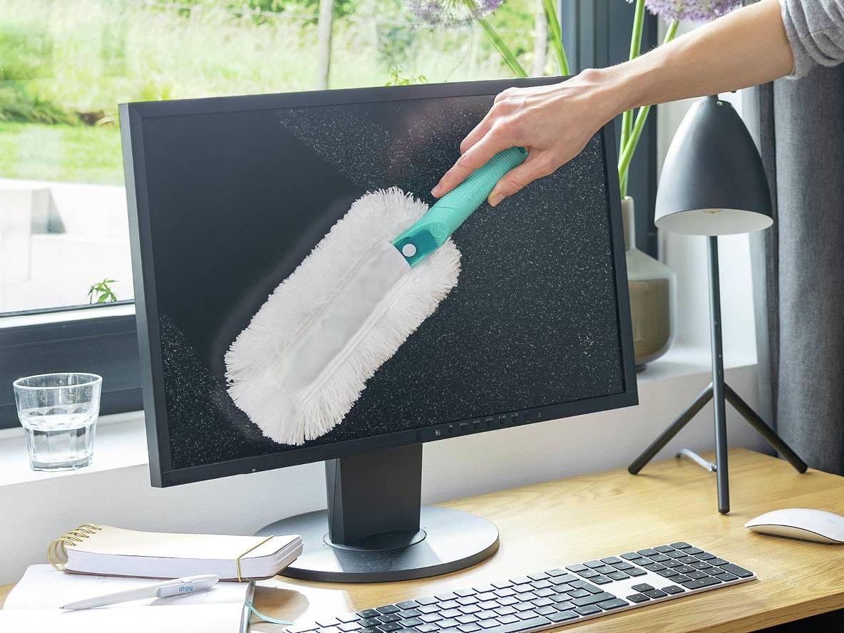 Hand wischt Staub von einem Computermonitor mit einem Staubwedel auf einem Schreibtisch