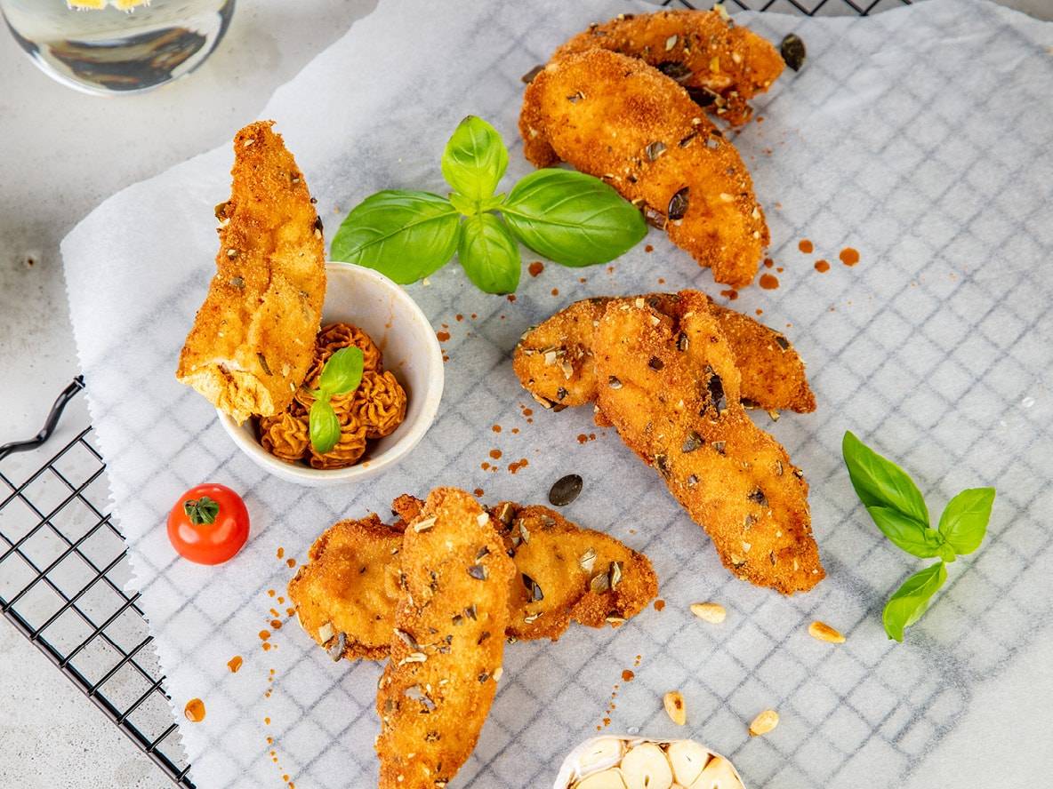 Knusprige Hähnchen-Nuggets mit Kernen, Basilikum und Dip auf Backpapier.