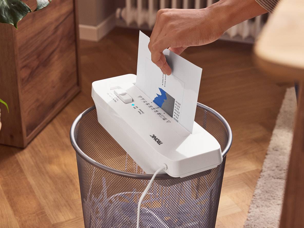 Eine Person schreddert ein Dokument mit einem Aktenvernichter, der auf einem Mülleimer steht.