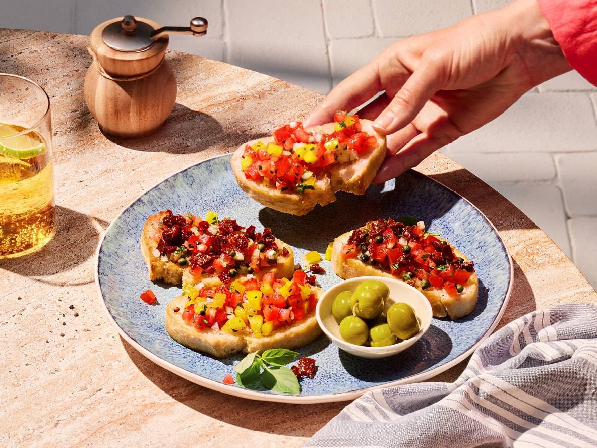 Une main attrape un morceau de bruschetta garni de tomates et de poivrons coupés en dés sur une assiette bleue.