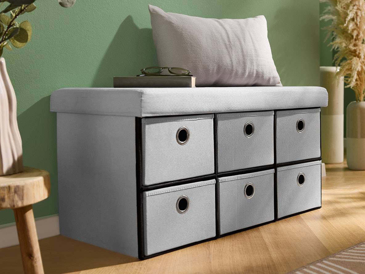 Graue Aufbewahrungsbank mit sechs Schubladen, Kissen und Brille vor grüner Wand.
