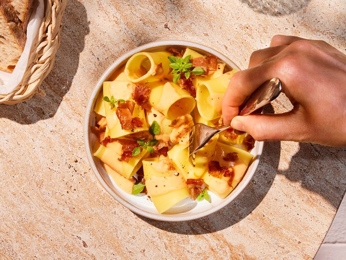 Eine Hand gabelt Nudeln mit Speck und Basilikum aus einer Schüssel auf einem Steintisch.
