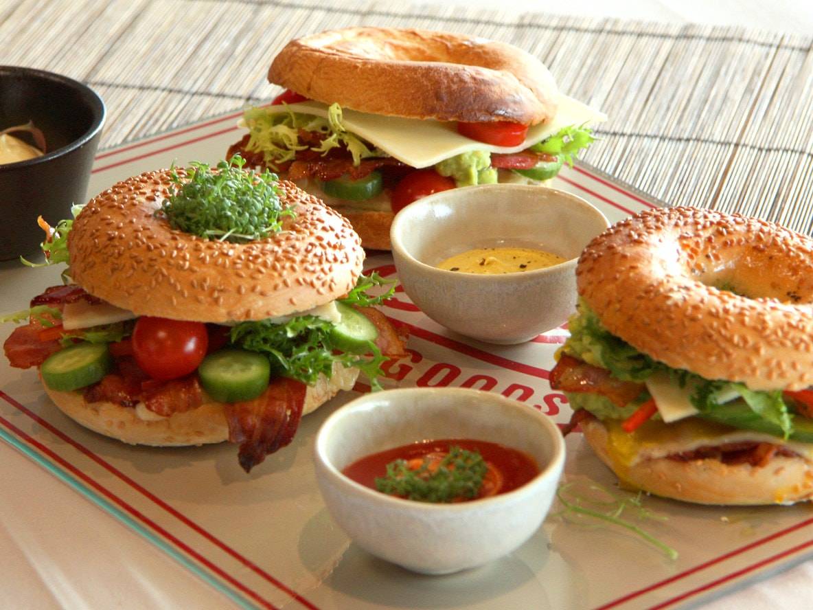 Drei Bagel-Sandwiches mit verschiedenen Füllungen und Dips auf einem Tablett.