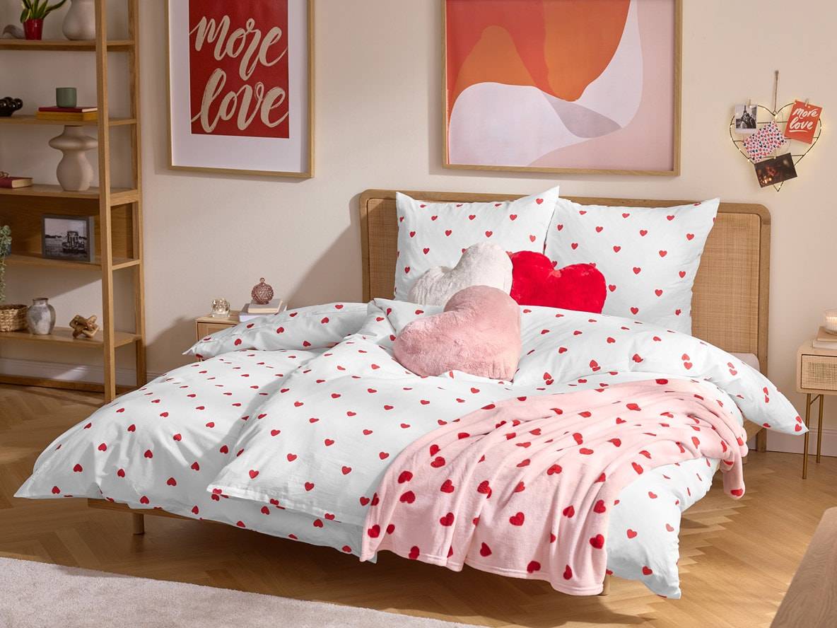 Parure de lit blanche à cœurs rouges, coussins en forme de cœur et couverture rose dans une chambre douillette.