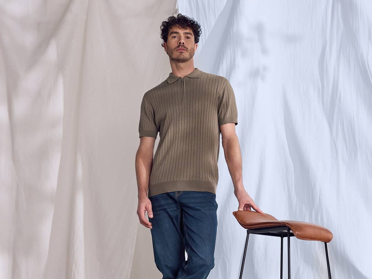 Homme en polo côtelé et jean, appuyé sur un tabouret marron.