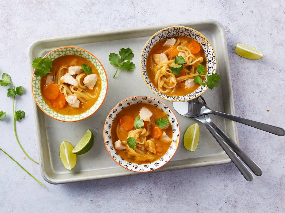 Trois bols de soupe de nouilles au poulet avec carottes, coriandre et quartiers de lime sur un plateau.