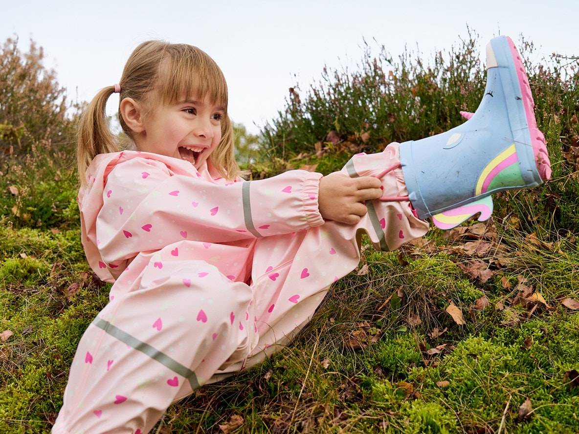 Enfant riant en imperméable rose à cœurs et bottes de pluie licorne.