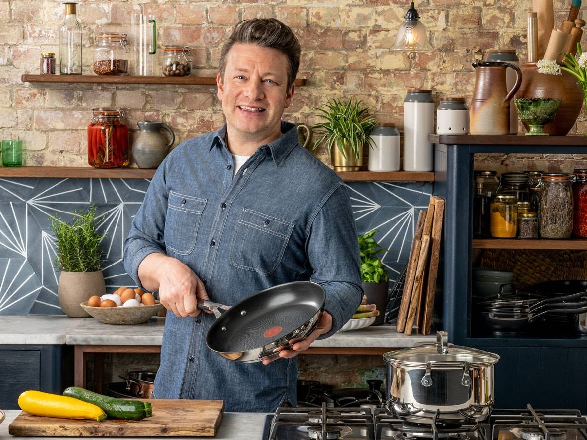 Ein lächelnder Koch hält eine Pfanne in einer rustikalen Küche mit Kochutensilien und Zutaten.