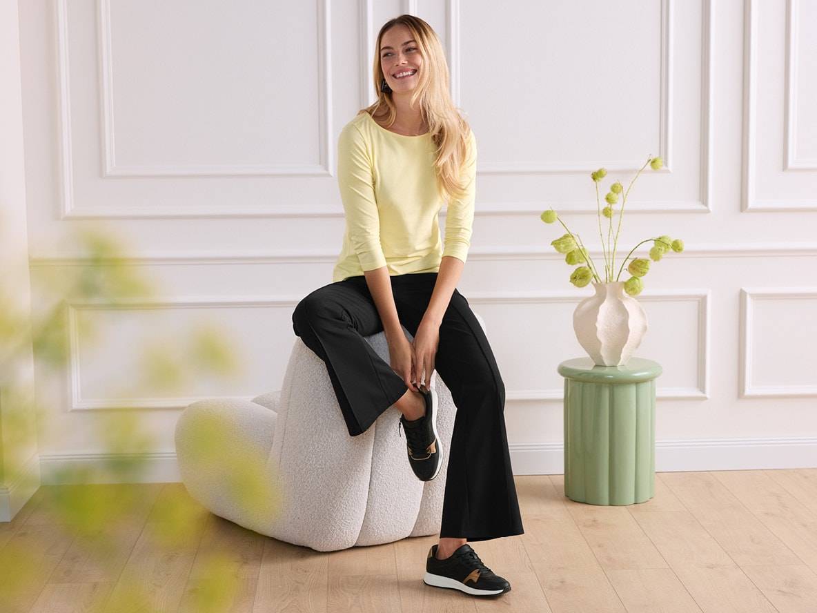 Eine lächelnde Frau in einem gelben Langarmshirt und schwarzen Hosen sitzt auf einem Sessel.