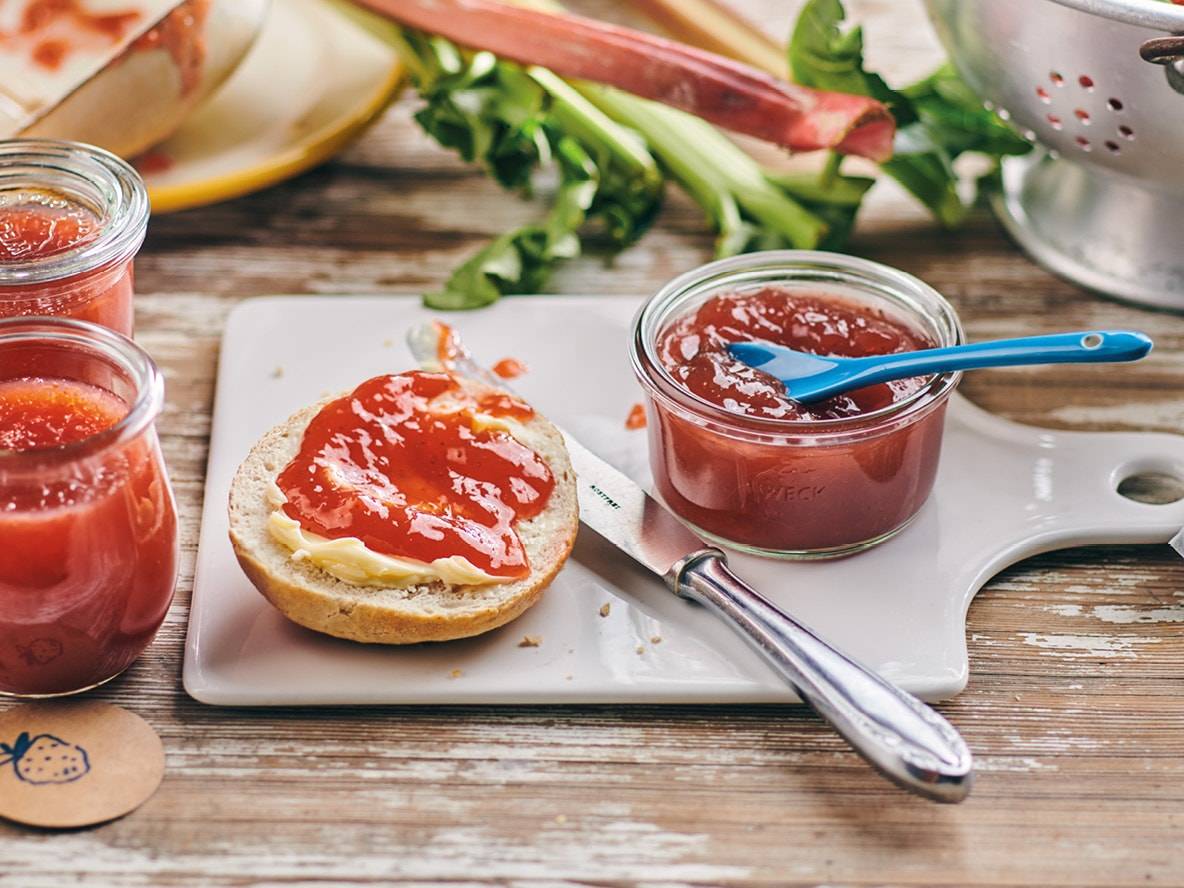 Rhabarber-Erdbeer-Marmelade auf einem Brötchen mit Butter, dazu Marmeladengläser und frischer Rhabarber.