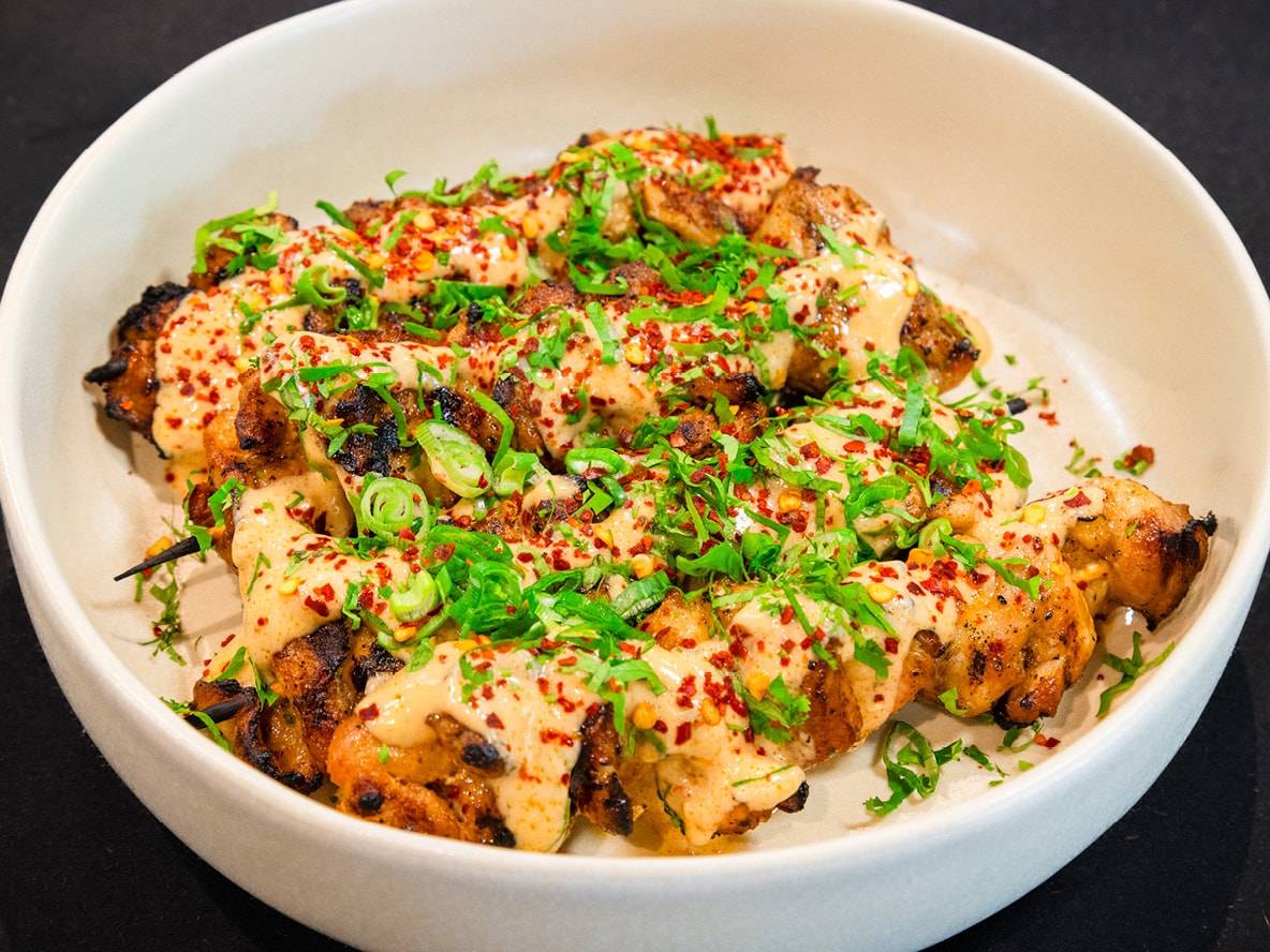 Spiedini di pollo grigliati con salsa cremosa, erbe fresche e scaglie di peperoncino in una ciotola bianca.