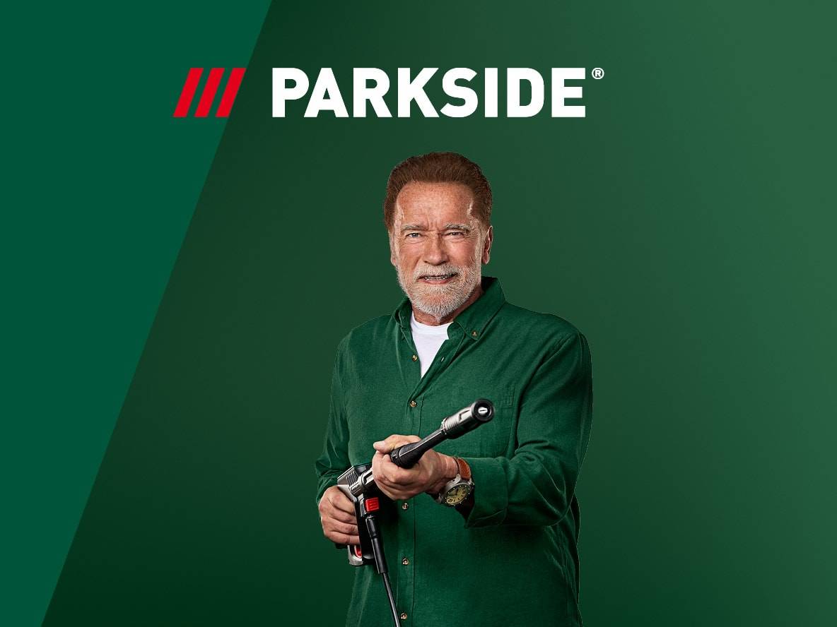 Arnold Schwarzenegger lächelt, hält ein Elektrowerkzeug vor grünem Hintergrund mit Logo.