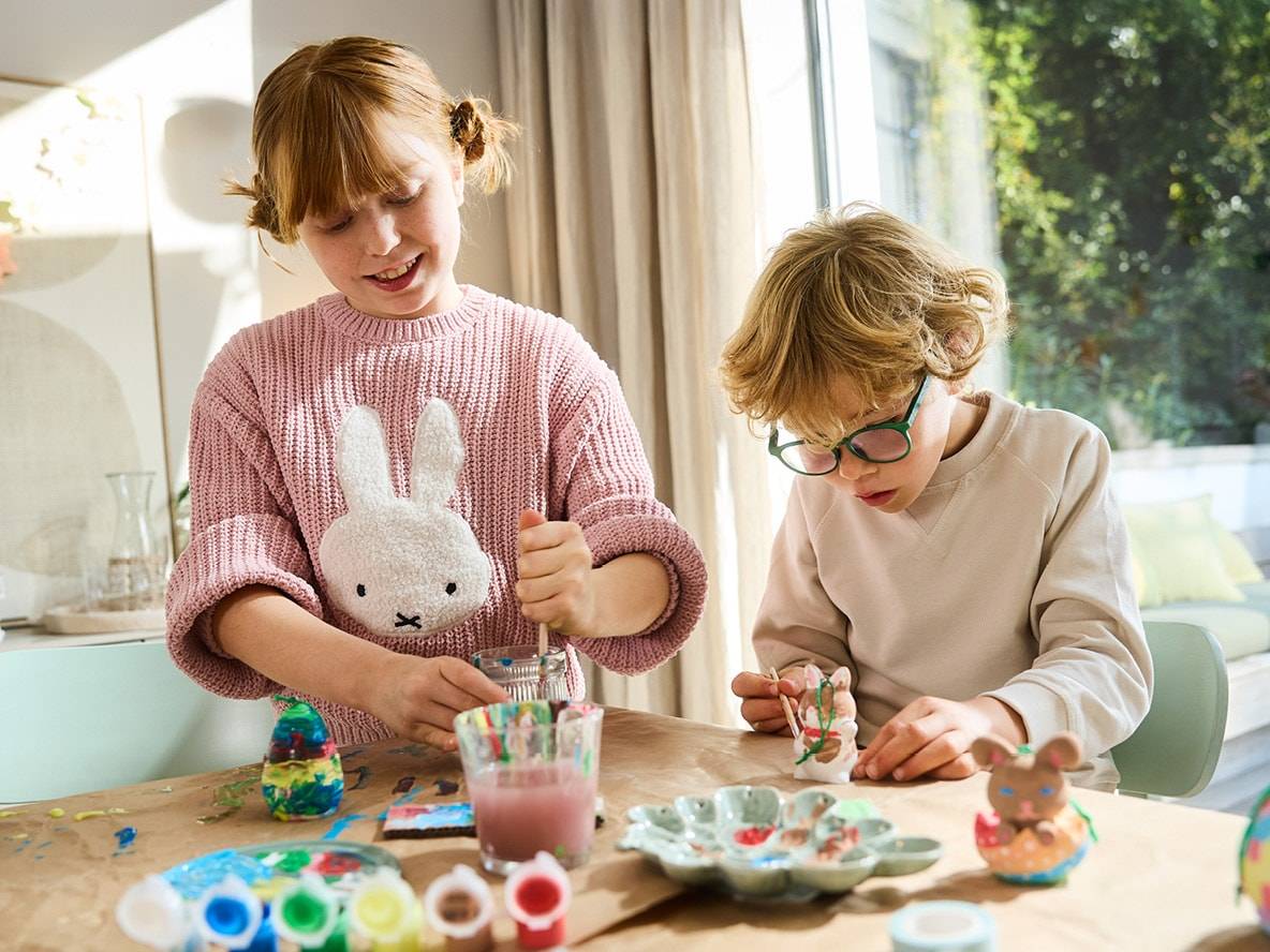 Zwei Kinder bemalen Osterhasenfiguren und Eier mit Farben und Pinseln.