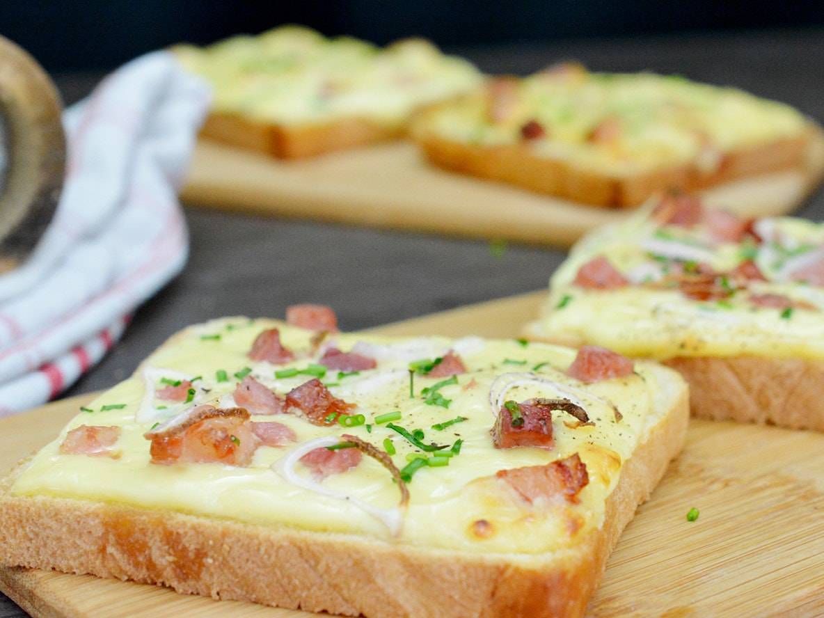 Gratinierte Toasts mit geschmolzenem Käse, Speck und Zwiebeln auf einem Holzbrett.